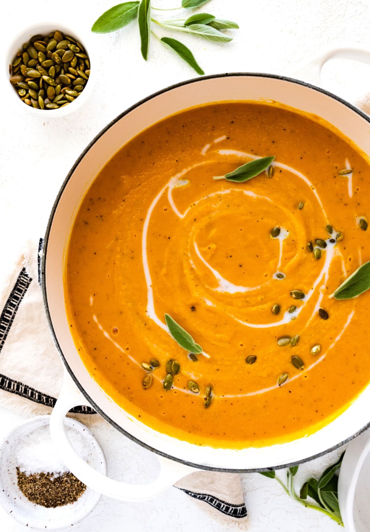 Butternut squash soup in a large pot garnished with coconut milk, pumpkin seeds, and fresh sage.