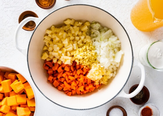 All the veggies and fruit used in the butternut squash soup in a large pot. This includes onion, garlic, carrot, ginger, and apple.