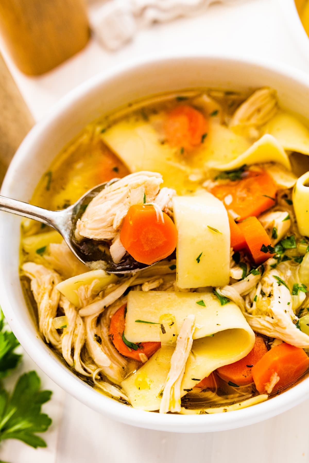 A spoon holding a bite size portion of chicken noodle soup over a bowl of the soup.