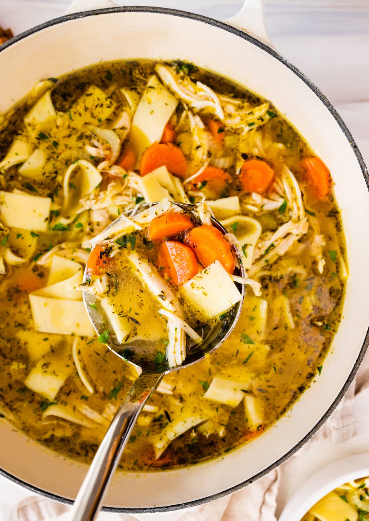 A metal ladle holding a portion of chicken noodle soup over a large pot of the soup.