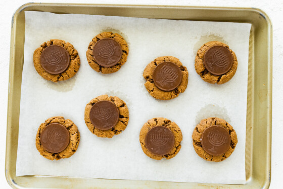 Multiple peanut butter blossoms on a baking sheet with a chocolate coin that depicts a menorah in the center of each cookie.