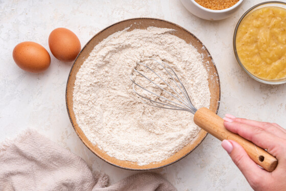 A woman's hand uses a metal whisk to whisk the flour in a bowl used for a banana bread.
