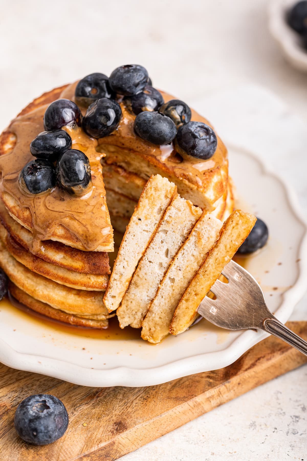 A stack of protein pancakes on a plate topped with fresh blueberries, nut butter, and maple syrup. A fork is taking a large bite-size portion from the stack of pancakes.