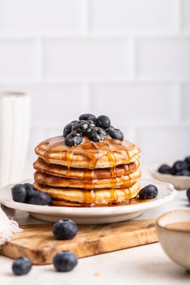 A stack of protein pancakes on a plate topped with fresh blueberries, nut butter, and maple syrup.