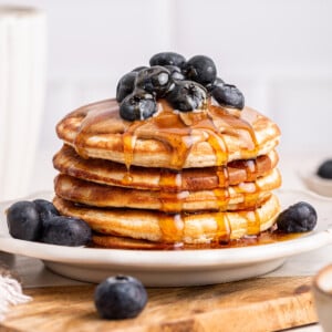 A stack of protein pancakes on a plate topped with fresh blueberries, nut butter, and maple syrup.