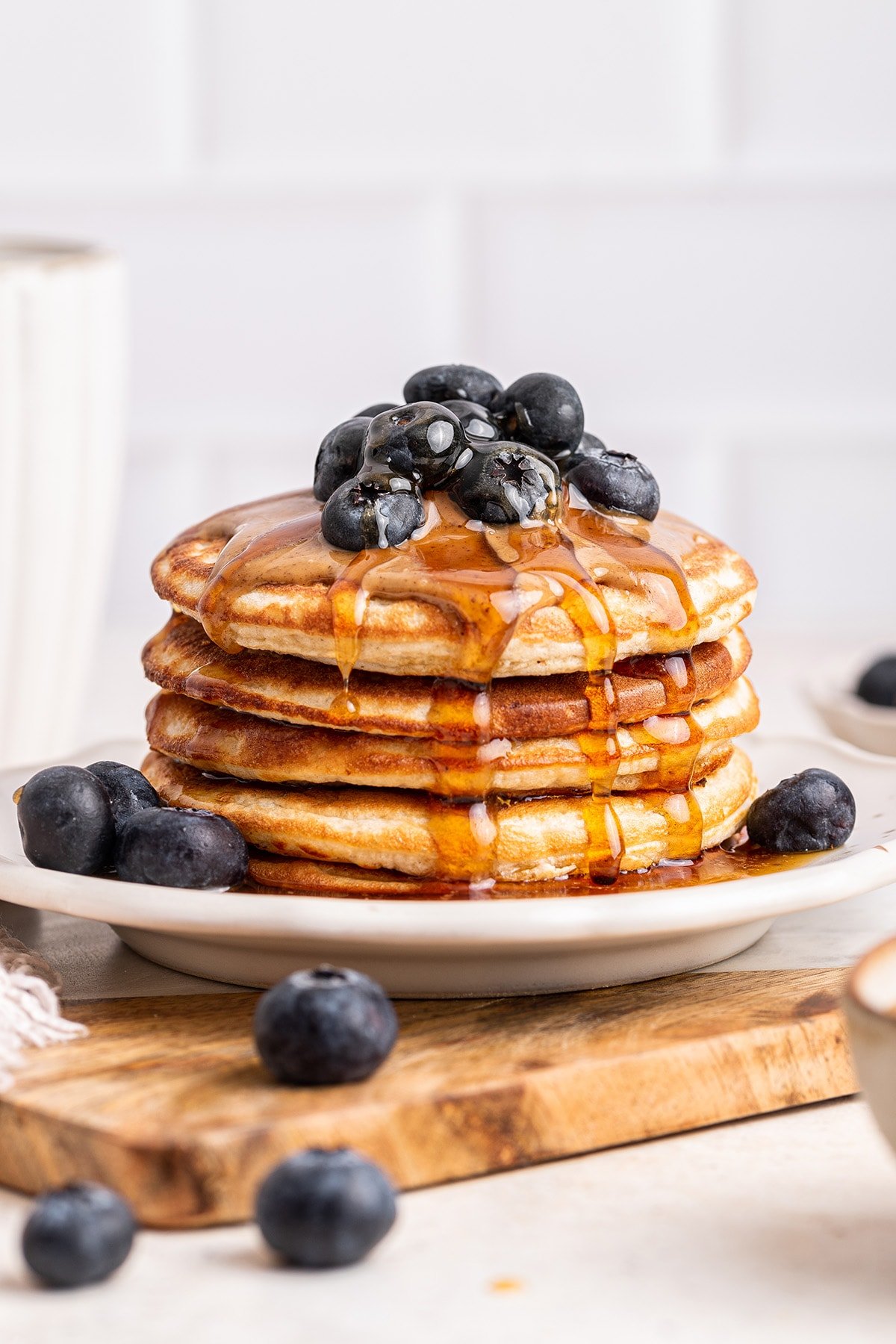 A stack of protein pancakes on a plate topped with fresh blueberries, nut butter, and maple syrup.