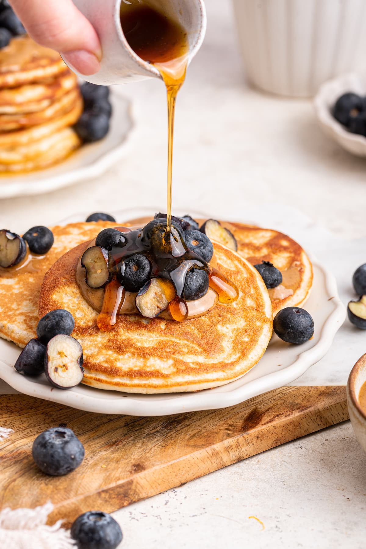 Maple syrup being drizzled onto protein pancakes that are on a plate and topped with fresh blueberries.
