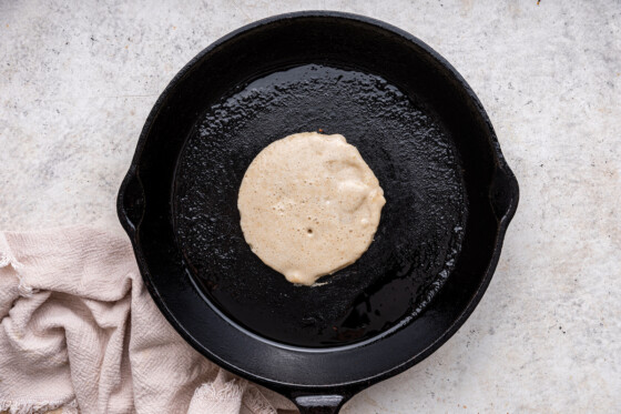Protein pancake batter in the shape of a pancake cooking in a cast iron skillet.