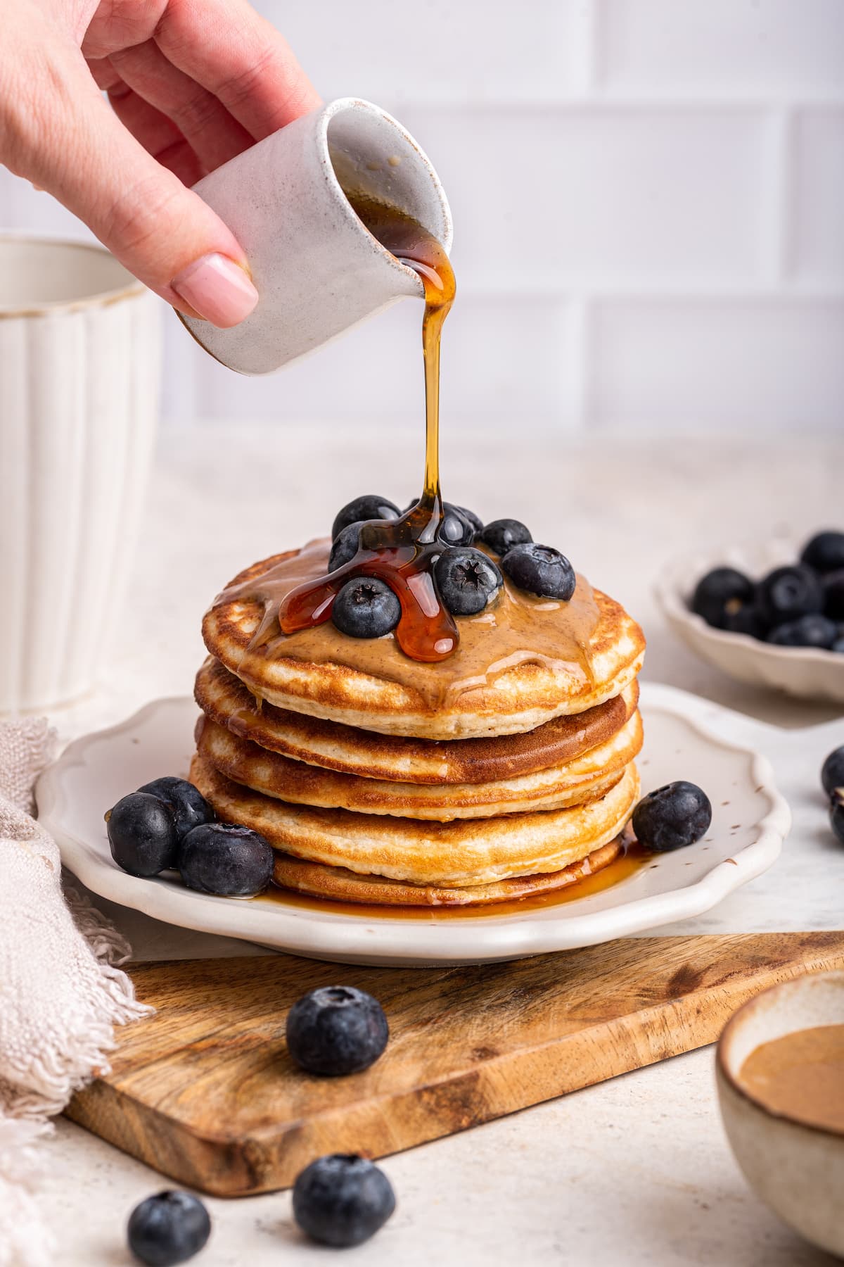 Maple syrup being poured over a stack of protein pancakes that are topped with fresh blueberries and nut butter.