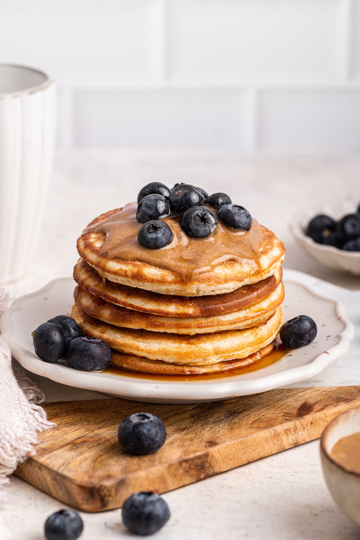 A stack of protein pancakes on a plate topped with fresh blueberries, nut butter, and maple syrup.