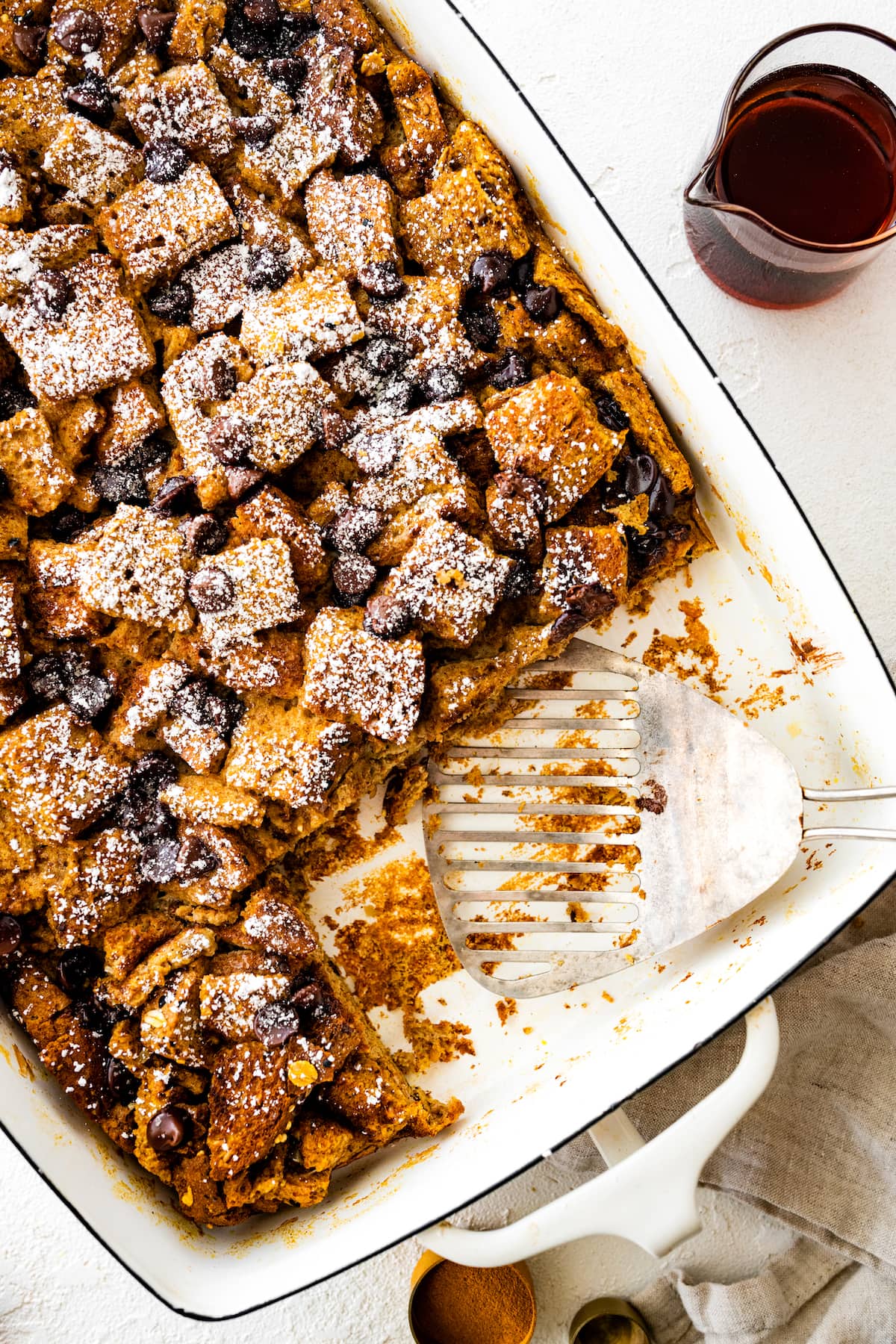Pumpkin French Toast Casserole in a square baking dish topped with powdered sugar and chocolate chips. Two pieces are missing from the dish where a metal spatula is instead.
