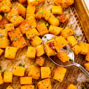 Seasoned and cubed roasted butternut squash on a baking tray with a metal spoon.