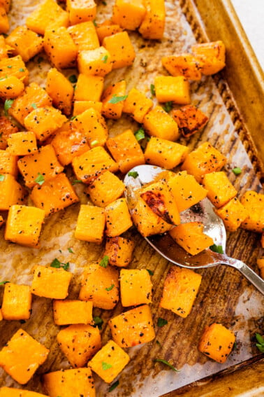 Seasoned and cubed roasted butternut squash on a baking tray with a metal spoon.