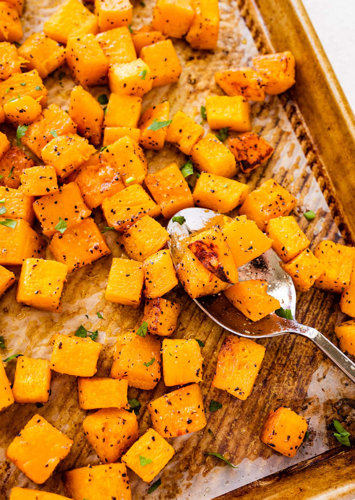 Seasoned and cubed roasted butternut squash on a baking tray with a metal spoon.