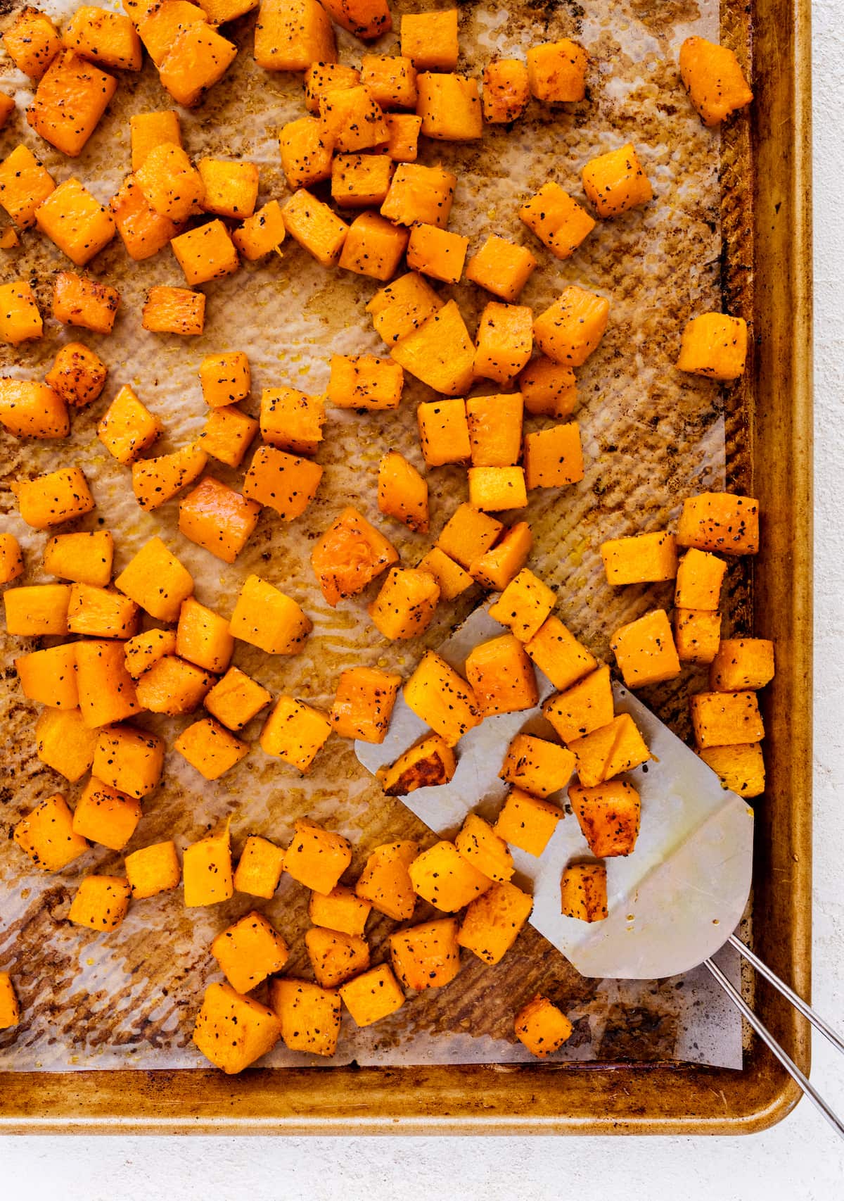 Seasoned and cubed roasted butternut squash on a baking tray with a metal spatula.