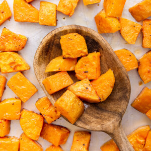 Cubed roasted sweet potatoes on a baking tray with a large wooden spoon holding a portion of these sweet potatoes.