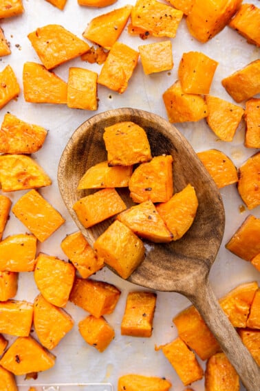 Cubed roasted sweet potatoes on a baking tray with a large wooden spoon holding a portion of these sweet potatoes.