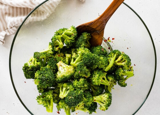 Seasoned broccoli in a large glass bowl with a large wooden spoon.