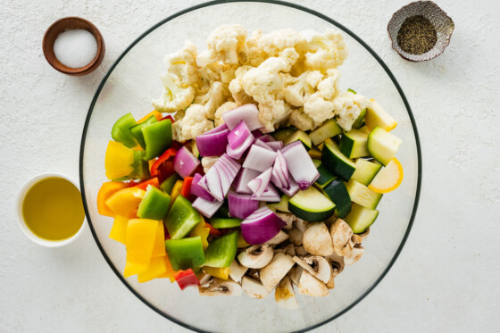 Raw vegetables in a large glass bowl. Vegetables include cauliflower, onion, zucchini, mushroom, and bell peppers.