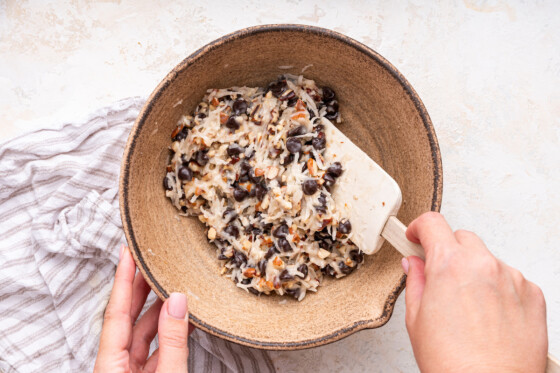 A woman's hand uses a silicone spatula to fold ingredients for chocolate coconut bars in a large mixing bowl.