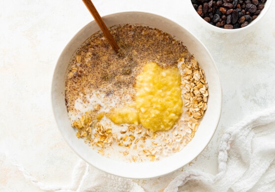 Mashed banana, milk, and vanilla extract added to the dry ingredients of cinnamon raisin baked oatmeal in a large mixing bowl.