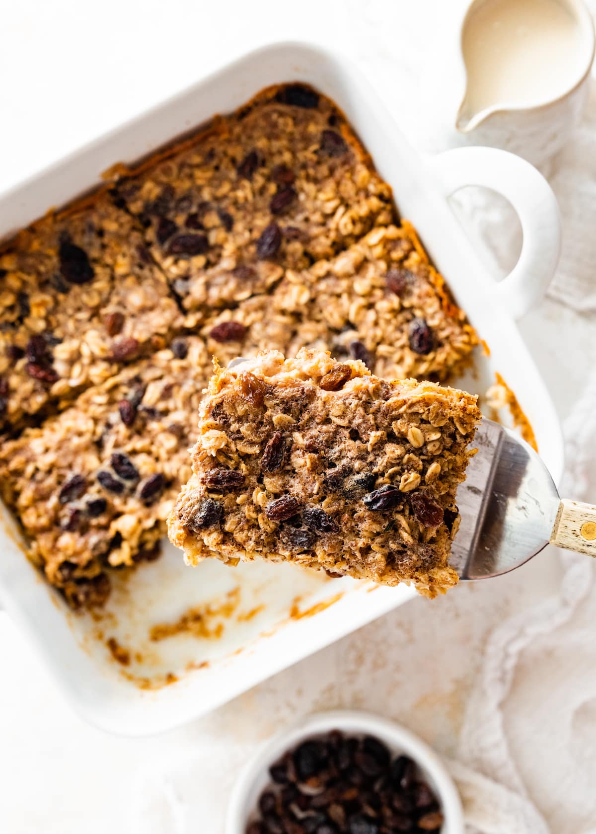 A metal spatula holding a serving of the cinnamon raisin baked oatmeal over a square baking dish with the rest of the baked oatmeal.