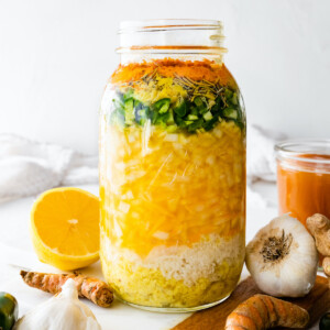 A large mason jar filled with all the ingredients used for a fire cider. Ingredients include horseradish, ginger, garlic, onions, cayenne, apple cider vinegar, and honey.