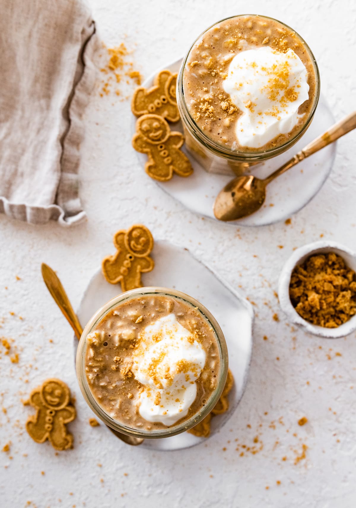 Two glass cups with gingerbread overnight oats topped with a dollop of whipped cream. The glass cups are on small plates with gingerbread men on them.