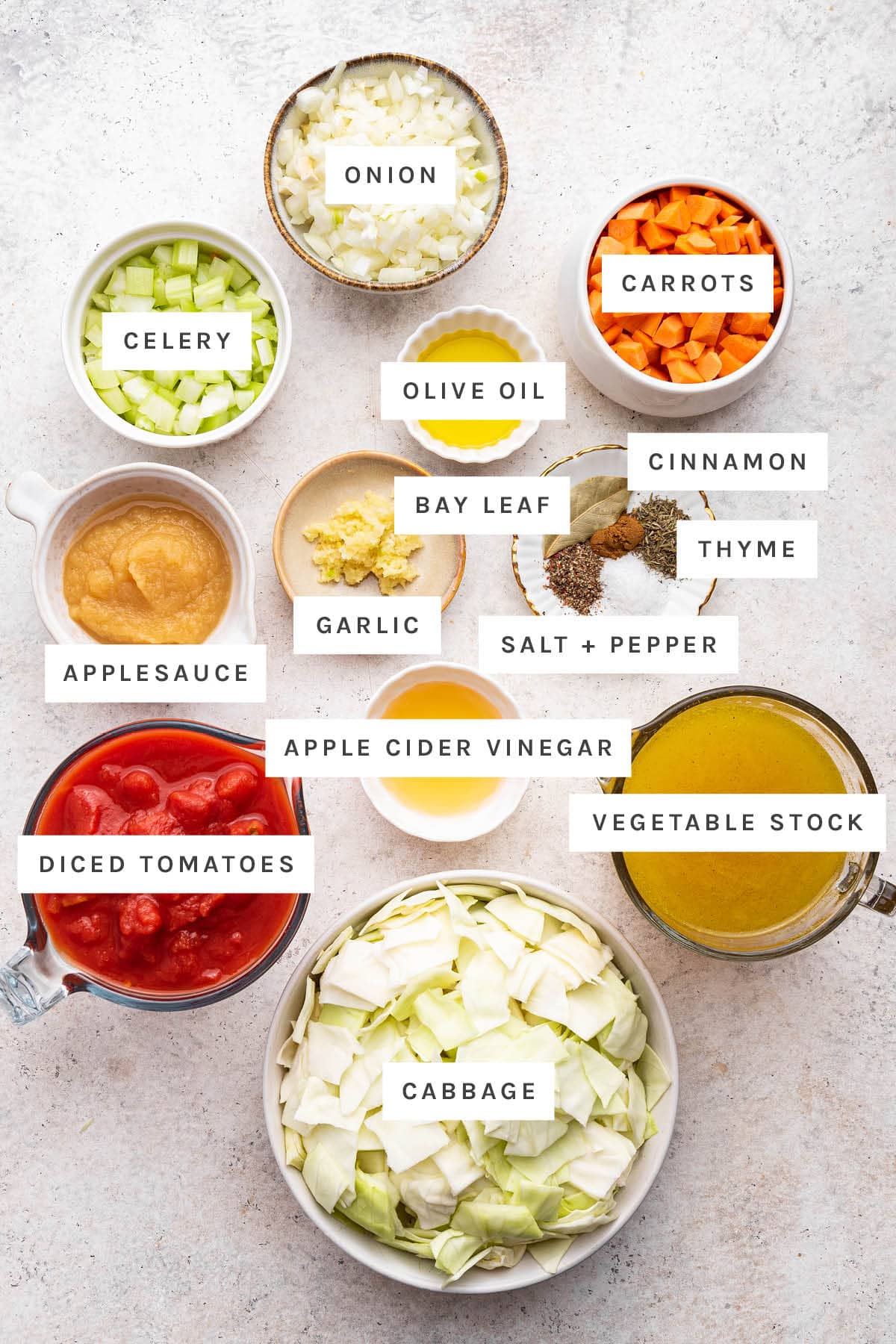 Ingredients measured out to make cabbage soup: applesauce, garlic, salt, pepper, bay leaf, thyme, diced tomatoes, onion, olive oil, cinnamon, vegetable stock, cabbage, carrots, celery and apple cider vinegar.