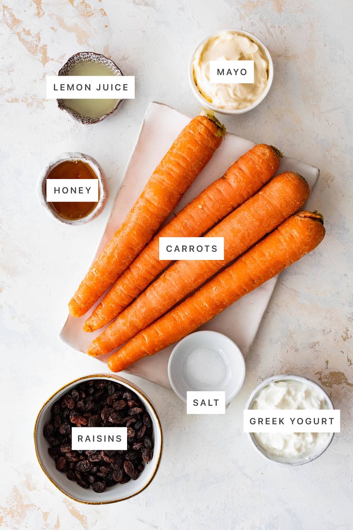 Ingredients measured out to make Carrot Raisin Salad: lemon juice, mayo, honey, carrots, salt, raisins and Greek yogurt.