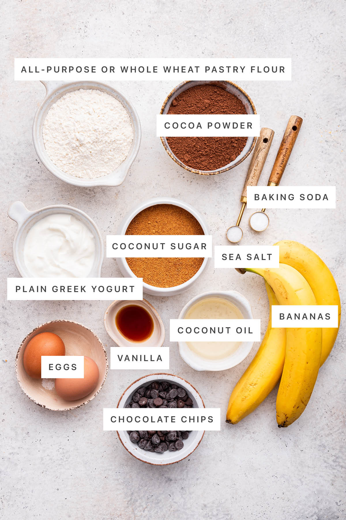 Ingredients measured out to make Chocolate Banana Bread: flour, cocoa powder, baking soda, coconut sugar, sea salt, Greek yogurt, eggs, vanilla, coconut oil, bananas and chocolate chips.