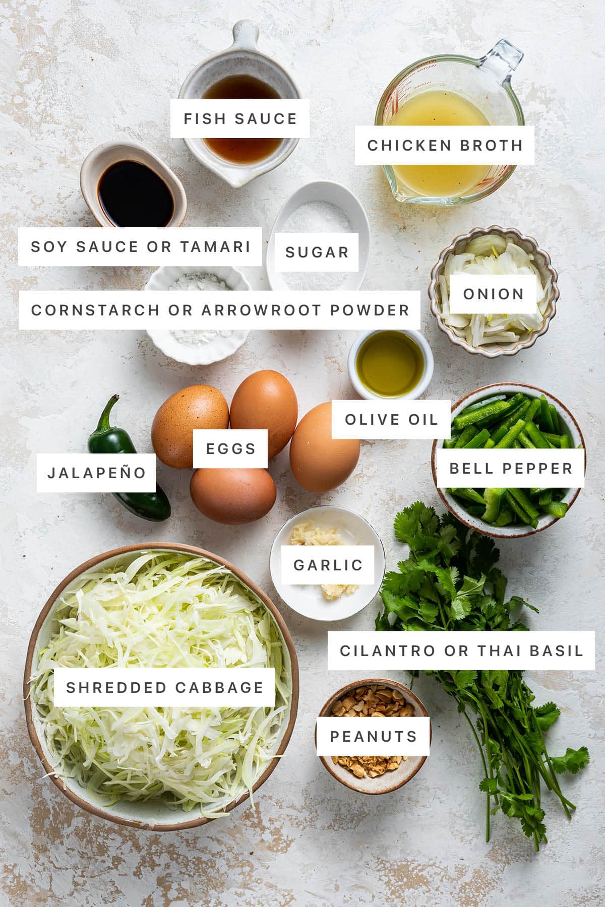 Ingredients measured out to make Healthy Pad Thai: fish sauce, chicken broth, soy sauce, sugar, onion, cornstarch, olive oil, eggs, jalapeño, bell pepper, garlic, cilantro, cabbage and peanuts.