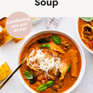 Bowl of lasagna soup topped with cheeses and fresh basil. Spoon is in the bowl. Bread, cheese and basil are beside the bowl.