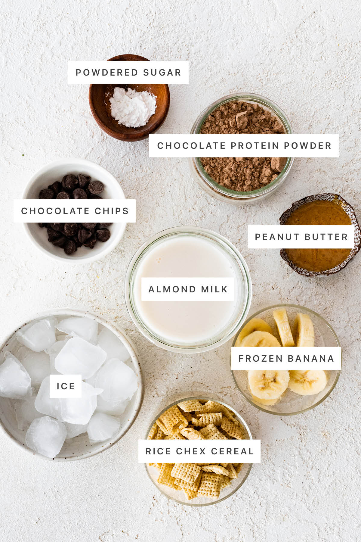Ingredients measured out to make Muddy Buddy Protein Shake: powdered sugar, chocolate protein powder, chocolate chips, peanut butter, almond milk, ice, frozen banana and Rice Chex cereal.