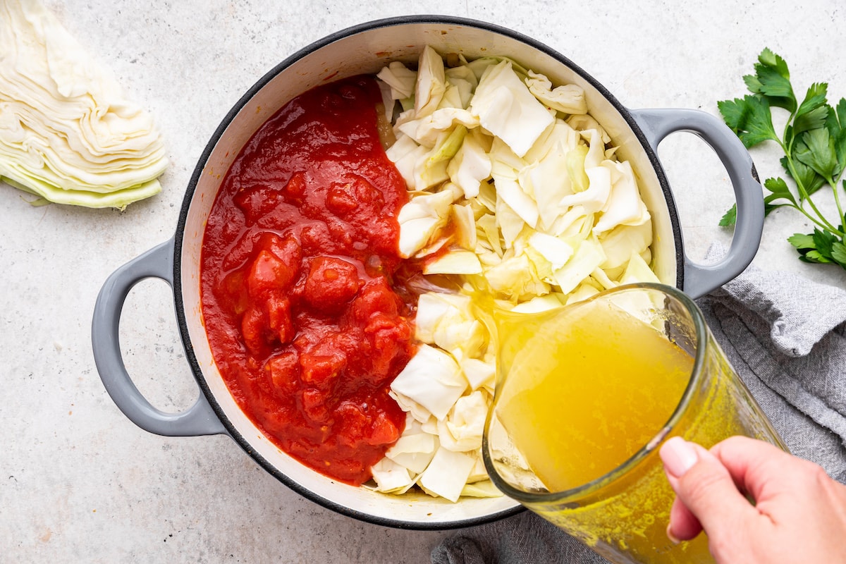 Broth being added to a large pot of vegetables, cabbage, and tomatoes.
