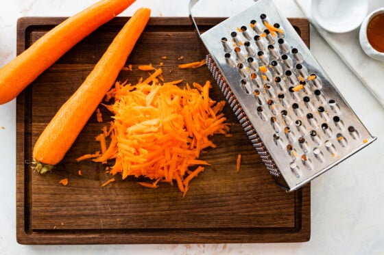Grated carrots on a wooden cutting board near two carrots and a grater.