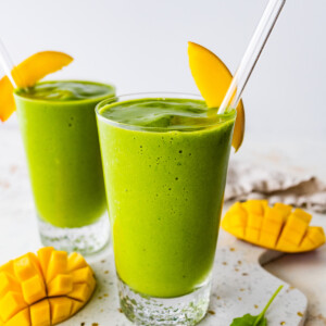 Two green smoothies in glasses with straws and fresh mango slices.