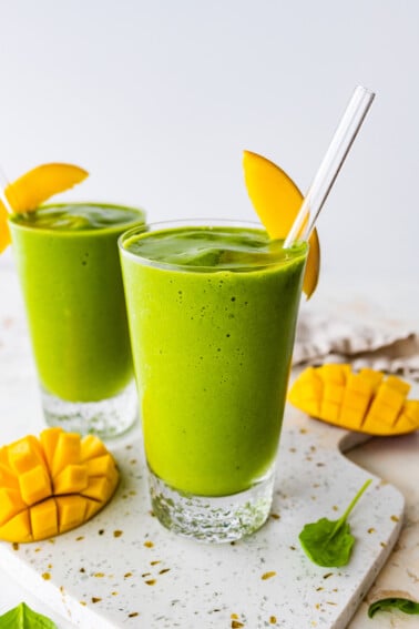 Two green smoothies in glasses with straws and fresh mango slices.