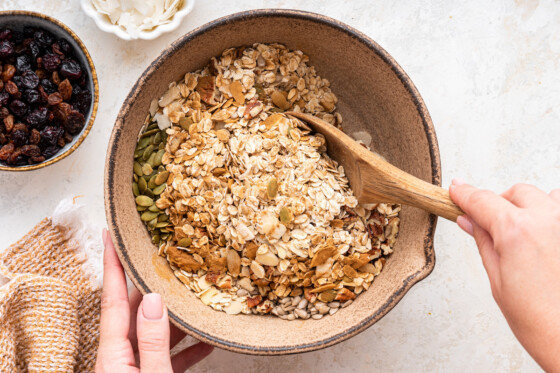 A woman's hand uses a wooden spoon to combine all the ingredients for muesli in a large mixing bowl.