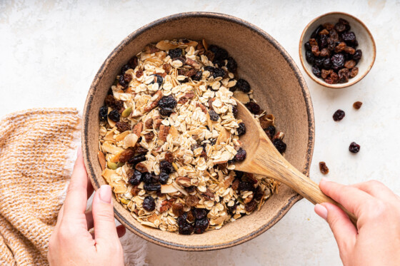 A woman's hand uses a wooden spoon to combine all the ingredients for muesli in a large mixing bowl.