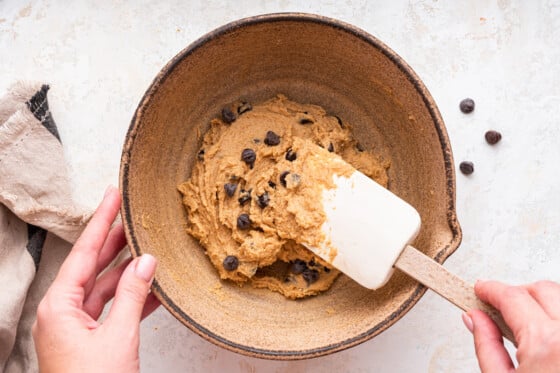A woman's hand uses a silicone spatula to mix in chocolate chips to the protein cookie dough.