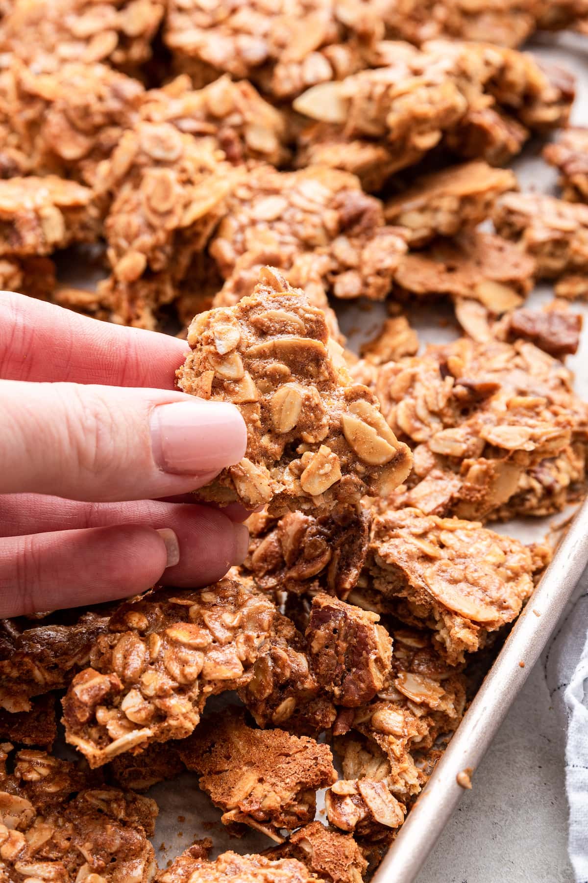 A woman's hand holds a piece of protein granola over a baking tray of the granola.
