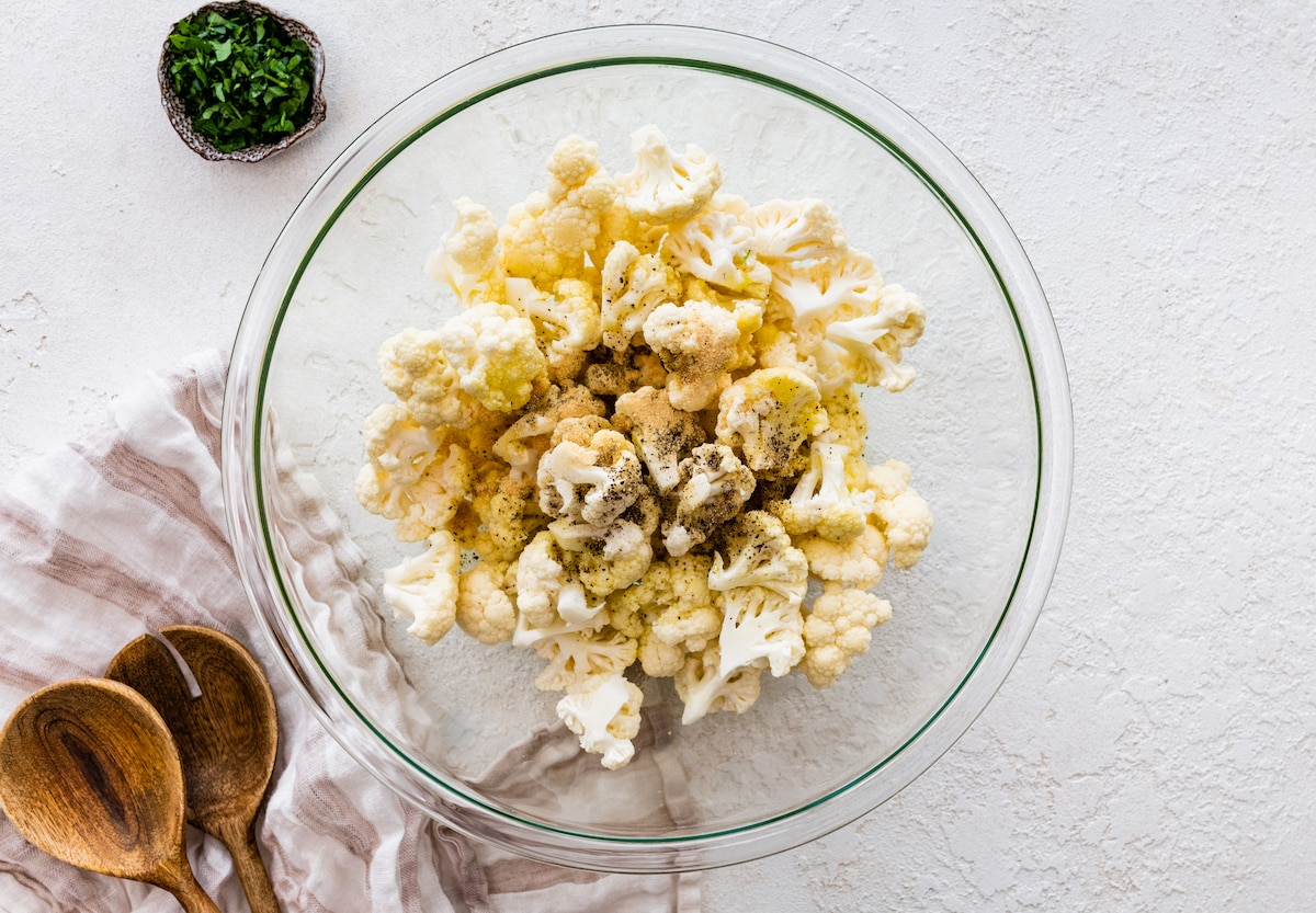 Raw cauliflower in a glass mixing bowl seasoned with salt, pepper and garlic powder.