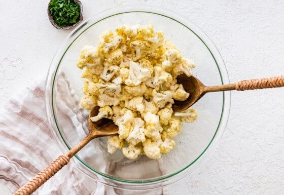 Raw cauliflower florets in a glass mixing bowl being tossed in oil and seasonings with two wooden spoons.