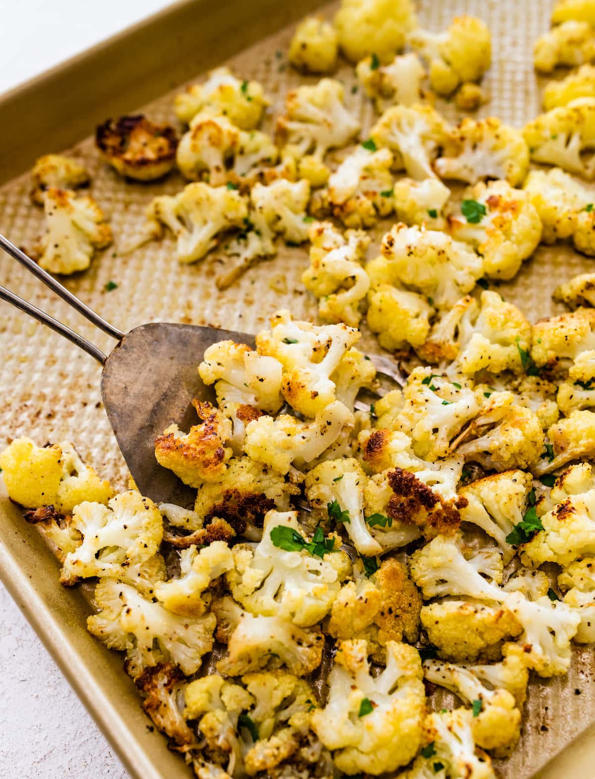 Roasted cauliflower on a baking sheet with a metal spatula scooping up some of the cauliflower.