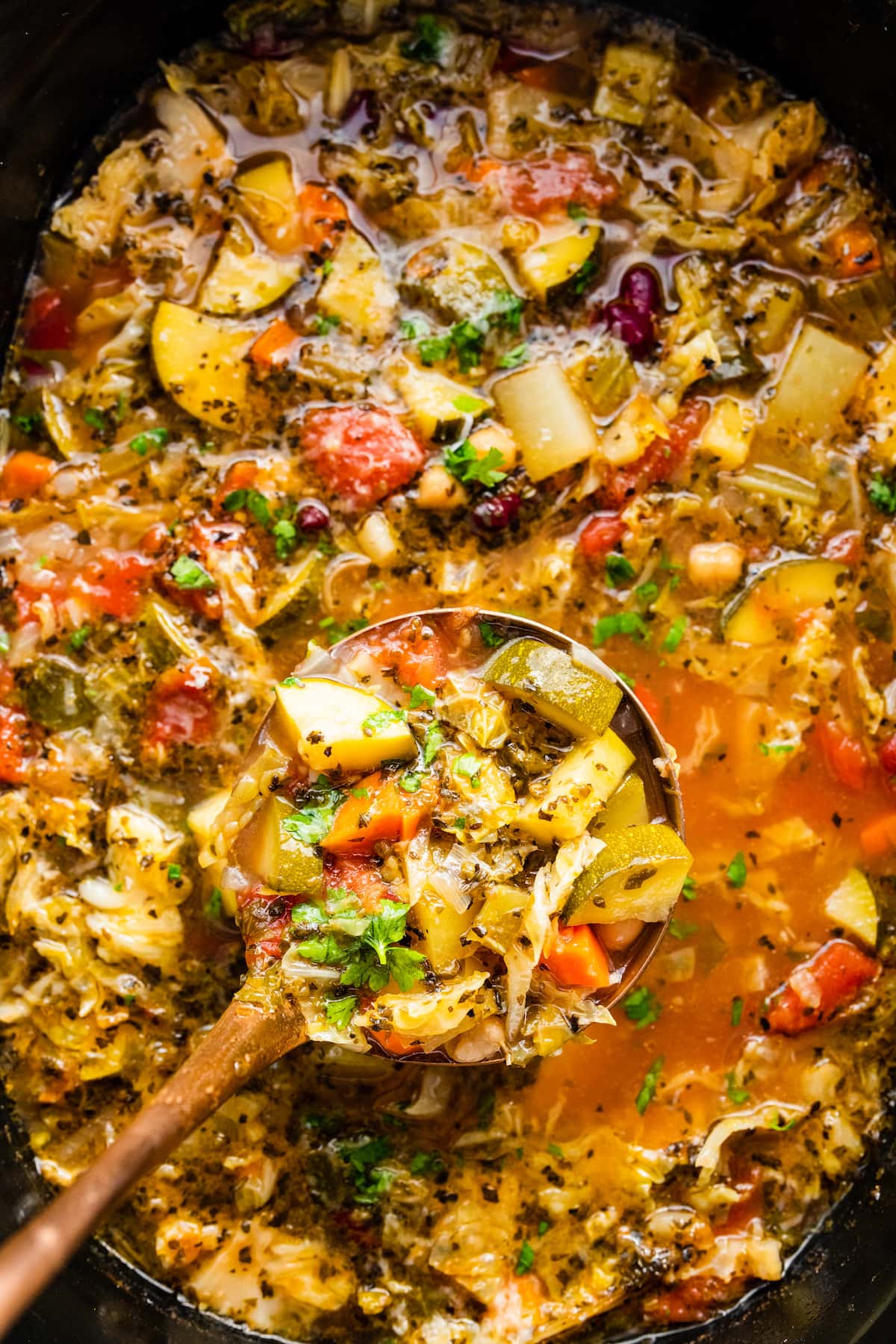 A large crock pot of minestrone soup with a ladle holding a portion of the soup.