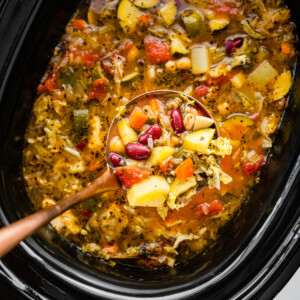 A ladle holding a portion of minestrone soup over a large crock pot of the soup.