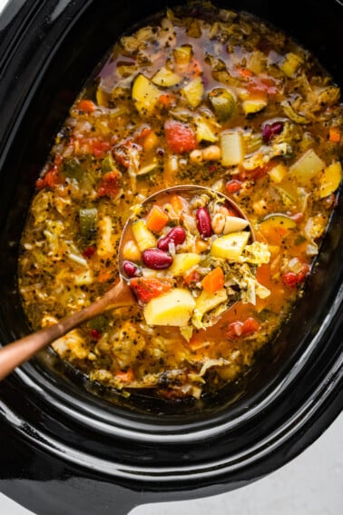 A ladle holding a portion of minestrone soup over a large crock pot of the soup.