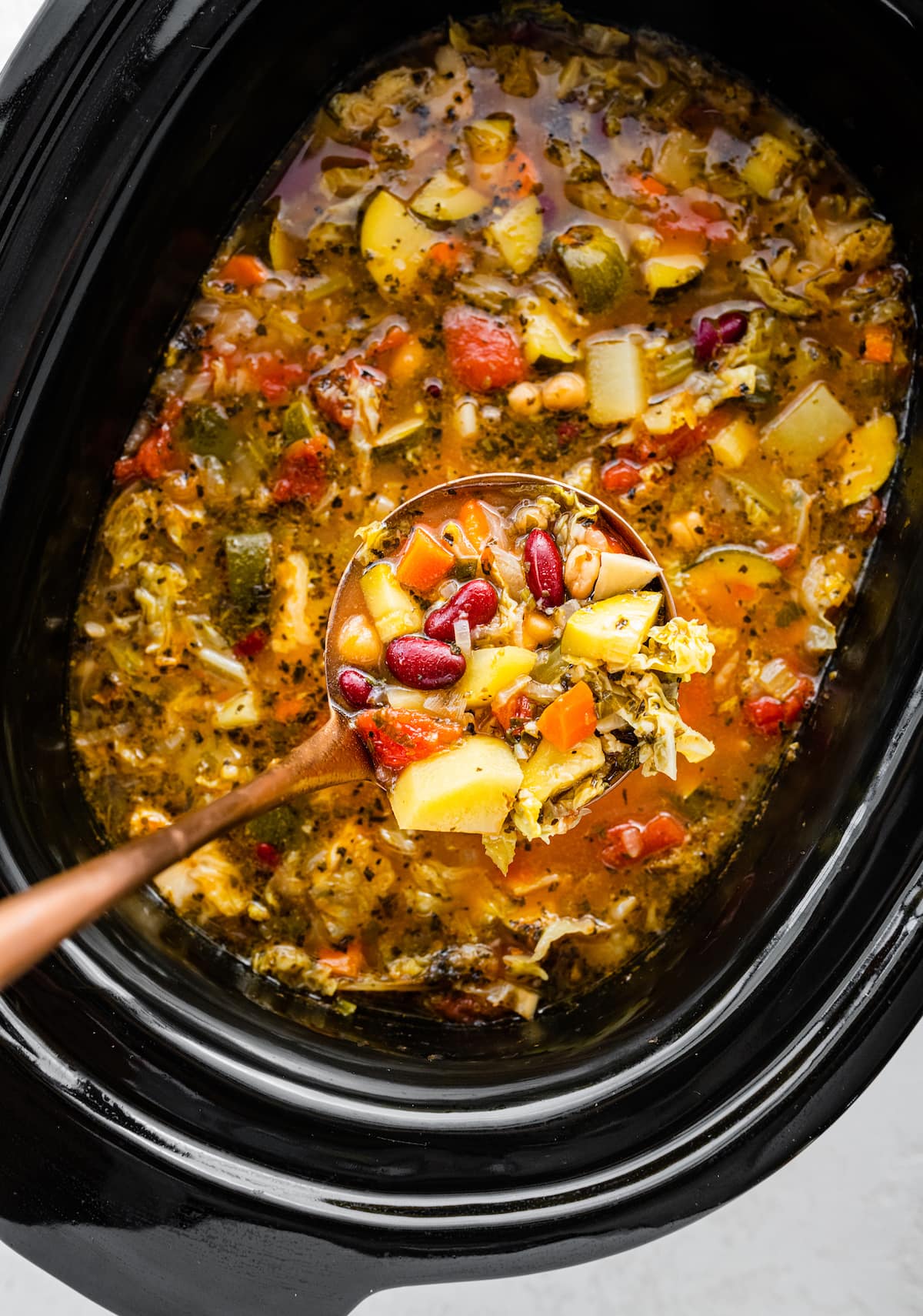 A ladle holding a portion of minestrone soup over a large crock pot of the soup.