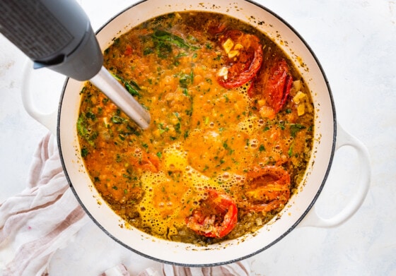 A hand blender being used to blend all the ingredients in a large pot to create a tomato basil soup.